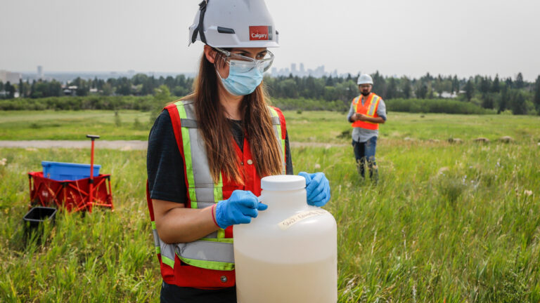 Une personne portant un gilet réfléchissant, un masque et un casque transporte une cruche blanche translucide contenant de l’eau jaune.