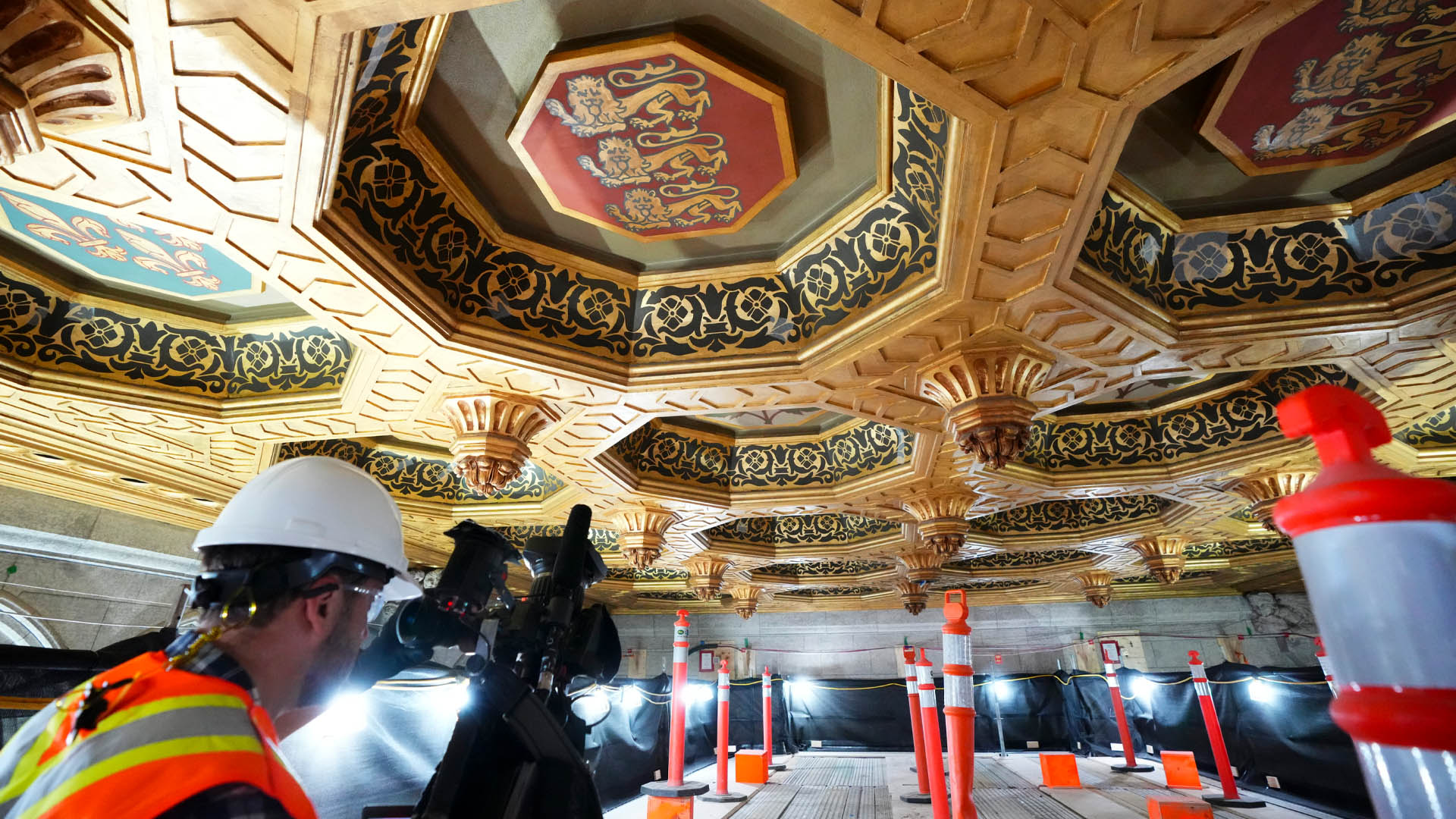 A worker in a reflective vest and hardhat stands on scaffolding just below the gilded Senate ceiling with red emblem featuring three lions who prepresent Canada’s historic ties to England.