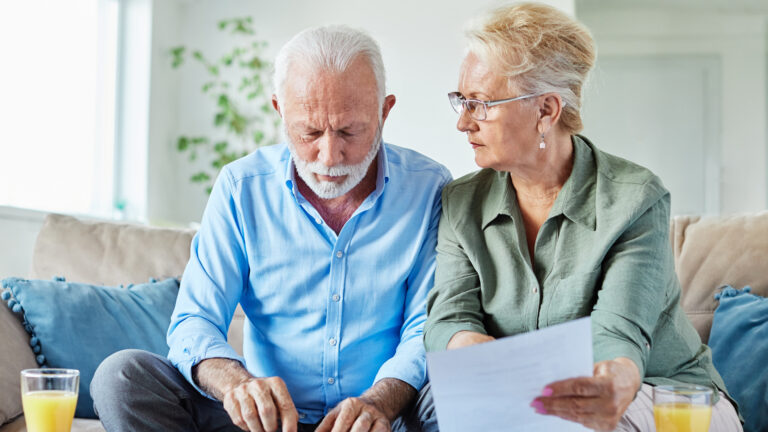 An older white man and woman sit on a sofa, looking through sheets of paper on a coffee table in front of them. He is using a calculator.