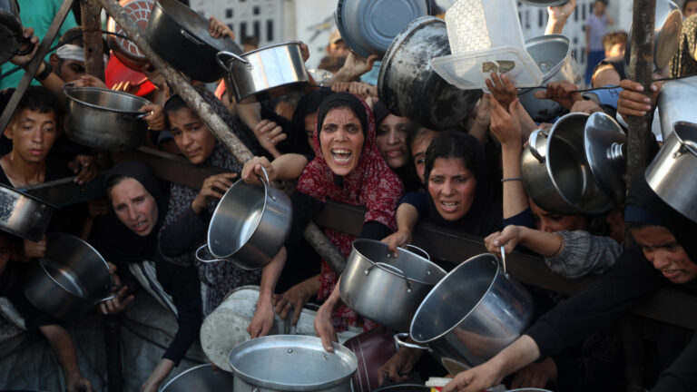 Women and youths reach out with metal pots at what appears to be a food distribution point. The food is not visible.