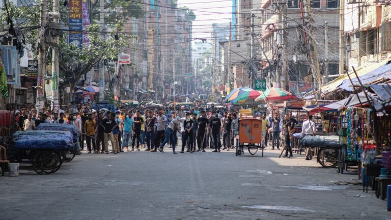 Hundreds of people stand on a busy city street that has a lot of overhead power lines crisscrossing above. The area in front of them has no traffic. Vendor stalls covered with tarps line the street.