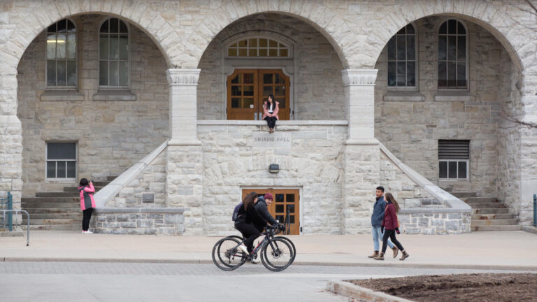 A grey stone university building with young people cycling and walking past while one person in a pink coat sits in an archway posing for a photo.