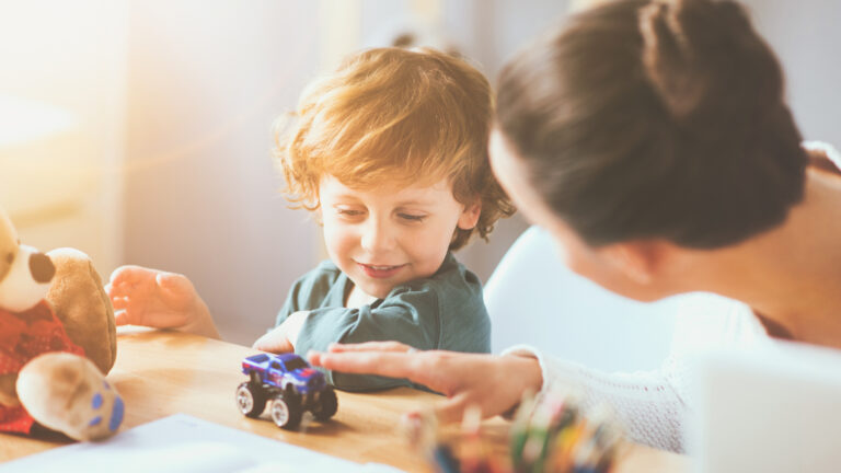 A woman speaks with a young boy sitting at a table with a tiny toy truck, a stuffed animal, colour pencils and sheets of paper in front of him.