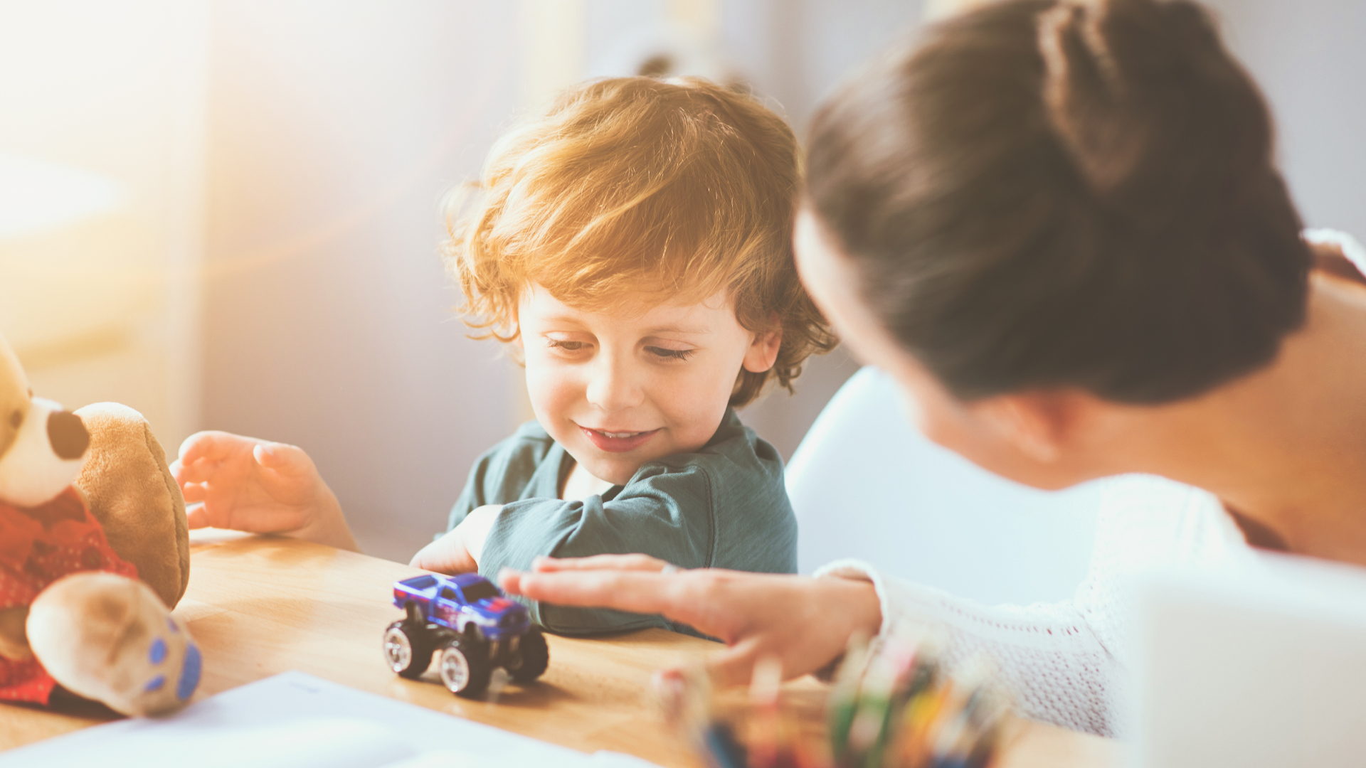 A woman speaks with a young boy sitting at a table with a tiny toy truck, a stuffed animal, colour pencils and sheets of paper in front of him.