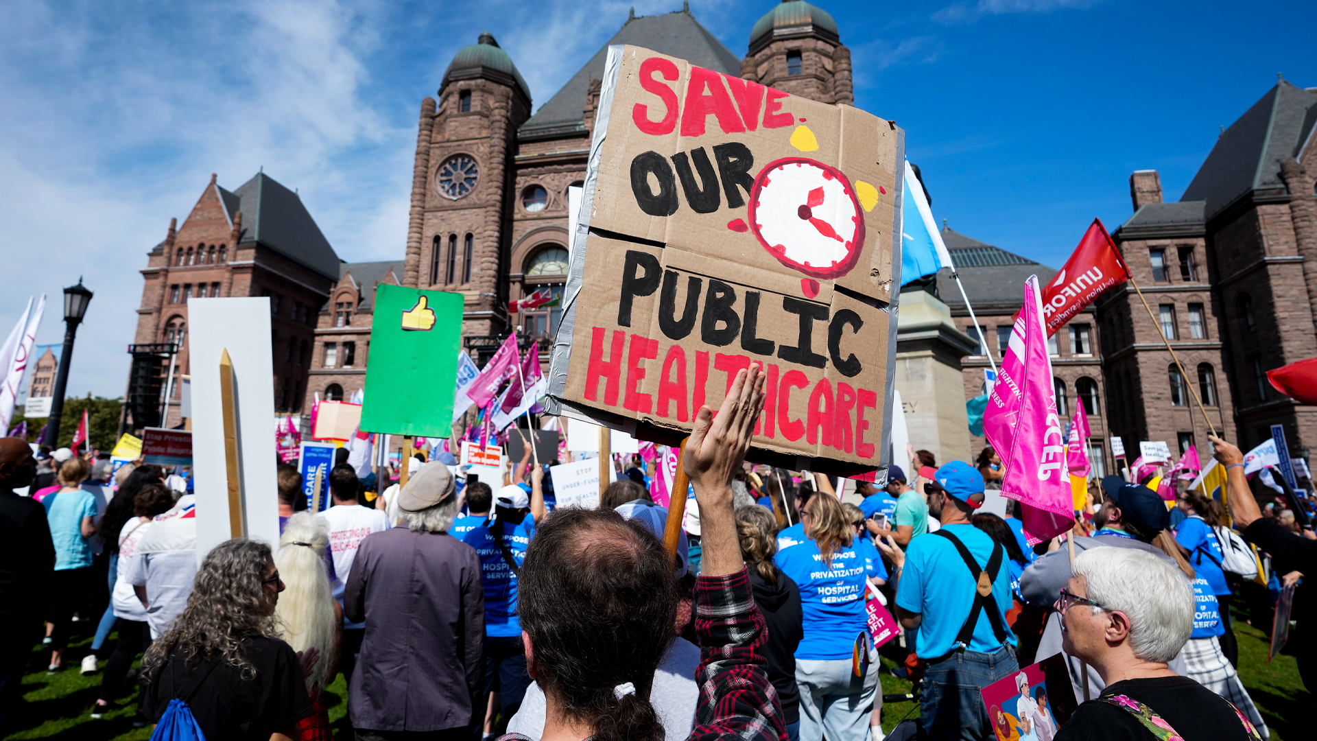 Dozens of people stand in front of the ornate, brown stone buildings. Many hold signs that say things like “Save our public healthcare.”