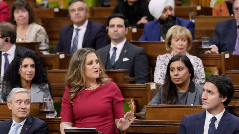 Chrystia Freeland, standing at her desk in a red dress in the House of Commons, speaks while Justin Trudeau and a dozen cabinet ministers look on.