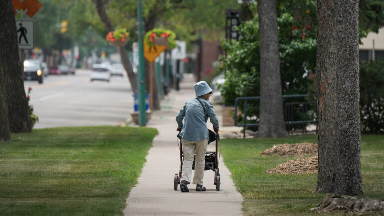The woman is wearing a blue hat and sunglasses and is walking away form the photographer on a leafy sidewalk.