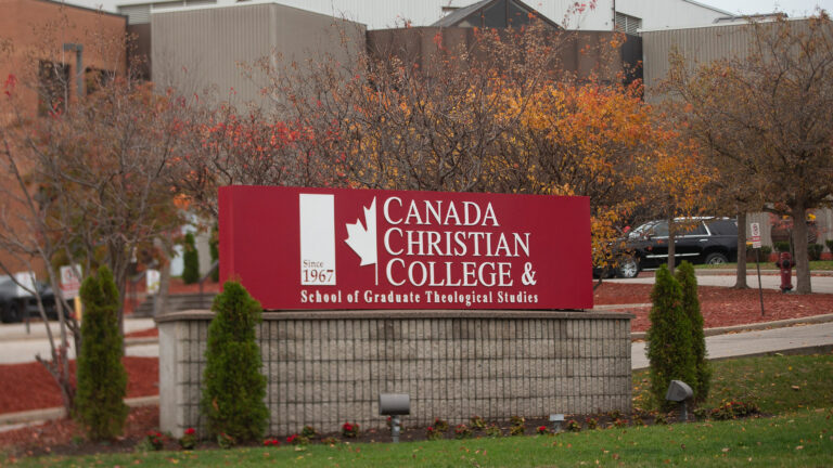 Brick, two-storey buildings stand behind a red and white sign for the college. Green grass, a couple small cedar hedges and several small trees with autumn-orange leaves surround the sign.