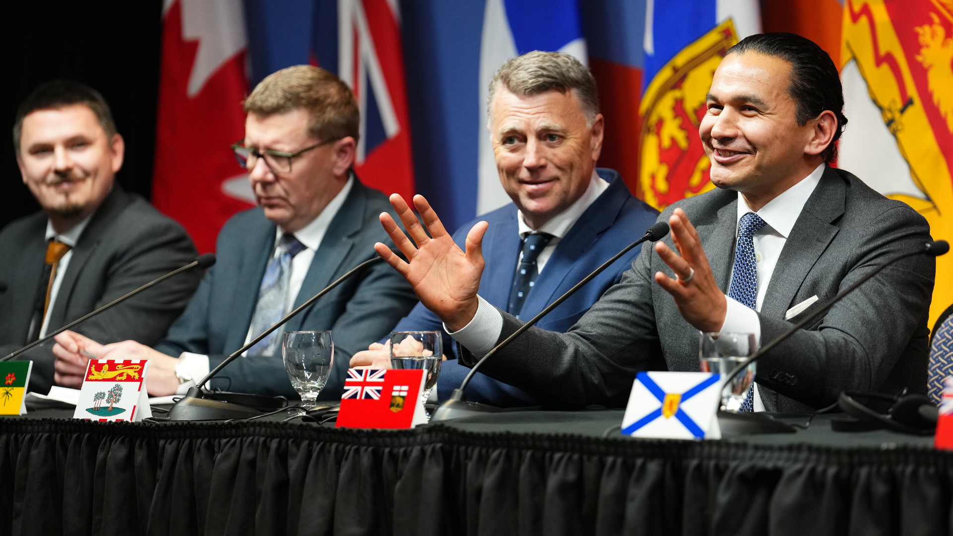 Le premier ministre du Manitoba, Wab Kinew, sourit en faisant des gestes avec ses mains, tandis que le premier ministre de l'Île-du-Prince-Édouard, Dennis King, assis à sa gauche, le regarde avec un sourire perplexe.
