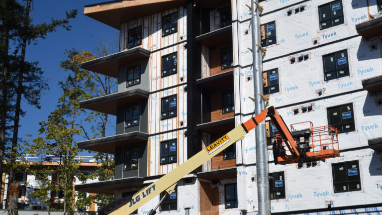 The building is six storeys high and is covered in a white Tyvek-branded membrane. A metal pole is attached to one wall. A crane lift sits idle next to it. The building has corner balconies and inset balconies. The window frames are black. Tall trees stand nearby.
