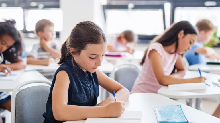 Two rows of children writing at desks in a classroom with large windows behind them.