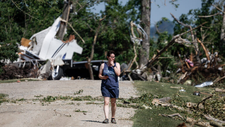 A woman holding her phone walks along a gravel rode with shattered trees and debris from homes and vehicles in the background.