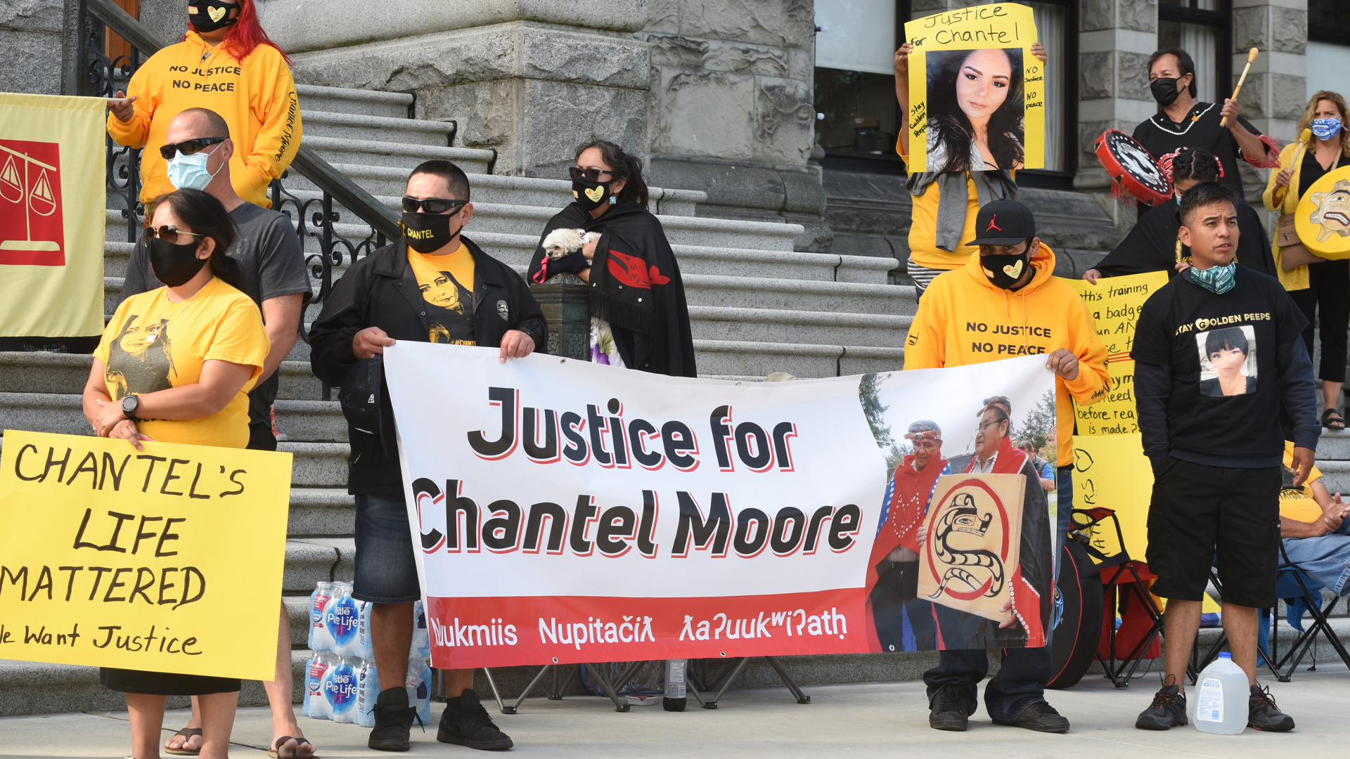At the base of a set of stone steps, about 10 people wear yellow hoodies and t-shirts that say “No justice, no peace” with photos of Chantel Moore on the fabric. Some people hold signs that say “Justice for Chantel Moore” and “Chantel’s life mattered; we want justice.”