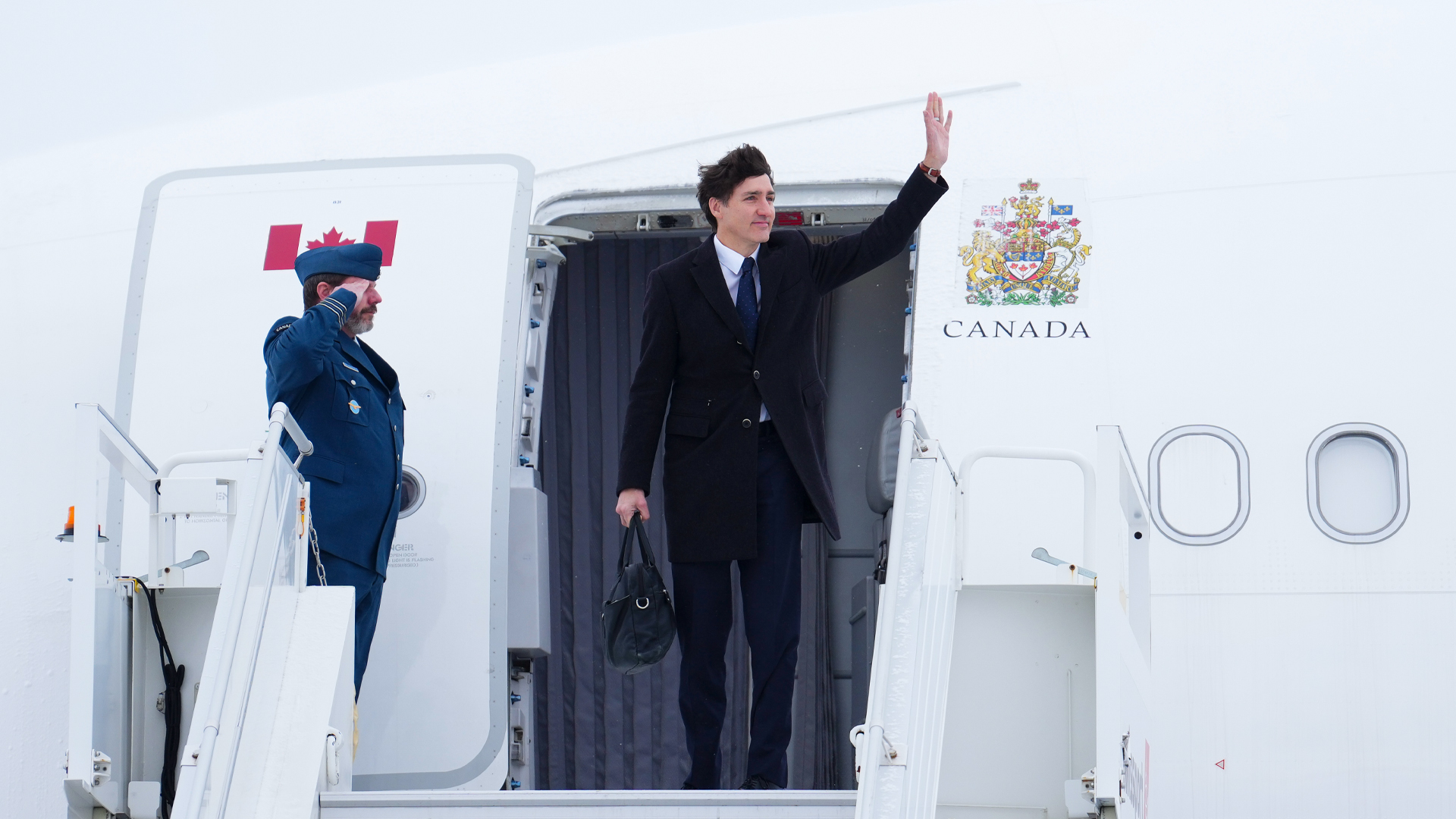 Justin Trudeau at the top of aircraft stairs, waving. To his right, a soldier salutes. To his left, Canada’s coat of arms is emblazoned on the side of the aircraft.