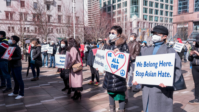 The people stand loosely in rows. They’re in winder coats and boots. Some hold signs saying “We belong here. Stop Asian Hate.” “We are all one.” “United against discrimination.” Everyone is wearing covid-masks.