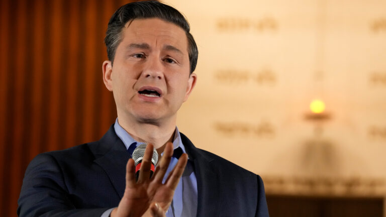 Pierre Poilievre, wearing a blue suit jacket and blue shirt without a tie, gestures with four fingers as he answers questions from reporters. The background is in soft focus but the event took place at a Shaarei Shomayim Congregation synagogue in Toronto.