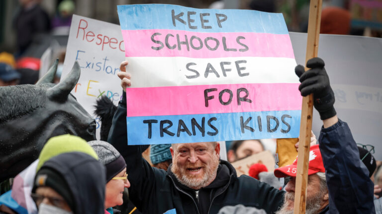 A bearded man holds a pink and blue sign saying “Keep schools safe for trans kids.”