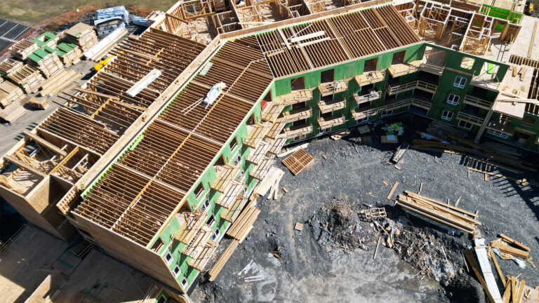 A four-storey building with is underway, with the top floor still in beginning stages and no roof yet. The balconies have their wooden beginnings, and the exterior wood walls are tinted green. Stacks of lumber sit on the far side of the building. Piles of construction debris are piled in front.