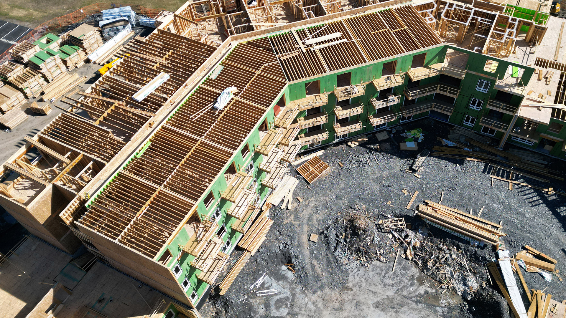 A four-storey building with is underway, with the top floor still in beginning stages and no roof yet. The balconies have their wooden beginnings, and the exterior wood walls are tinted green. Stacks of lumber sit on the far side of the building. Piles of construction debris are piled in front.