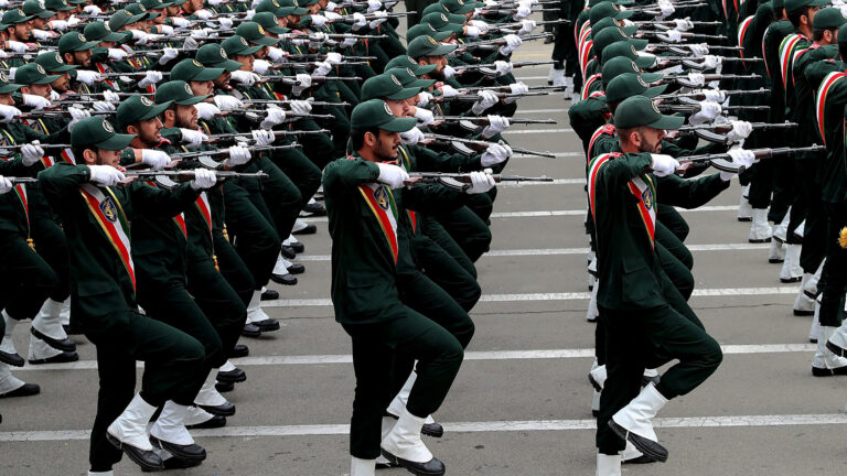 Hundreds of young men march in rows, dressed head-to-toe in dark green with white spats over their black boots and a red, white and blue sash draped across their chests. Wearing white gloves, each man is holding a long weapon at shoulder level.