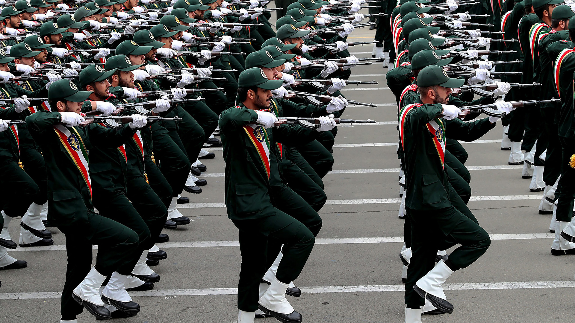 Hundreds of young men march in rows, dressed head-to-toe in dark green with white spats over their black boots and a red, white and blue sash draped across their chests. Wearing white gloves, each man is holding a long weapon at shoulder level.