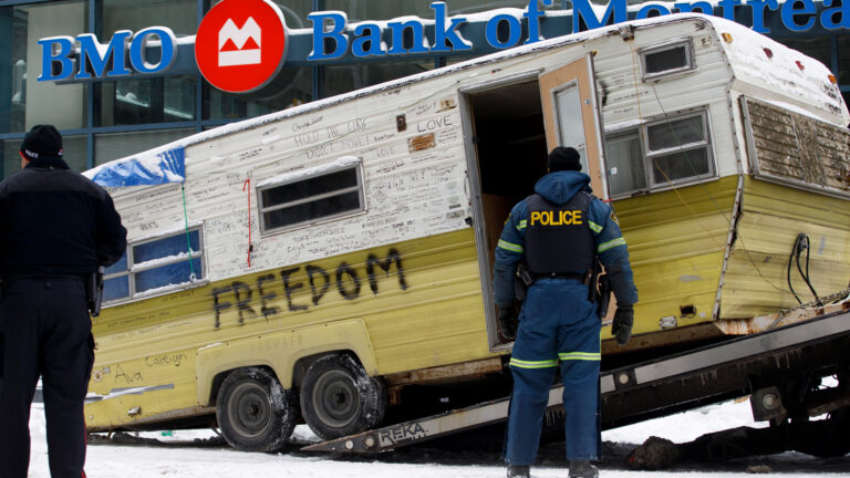 On a snow-covered street, a dingy-looking yellow and white camper covered with dozens of words like “freedom” is loaded onto a flatbed as two police officers supervise the operation.