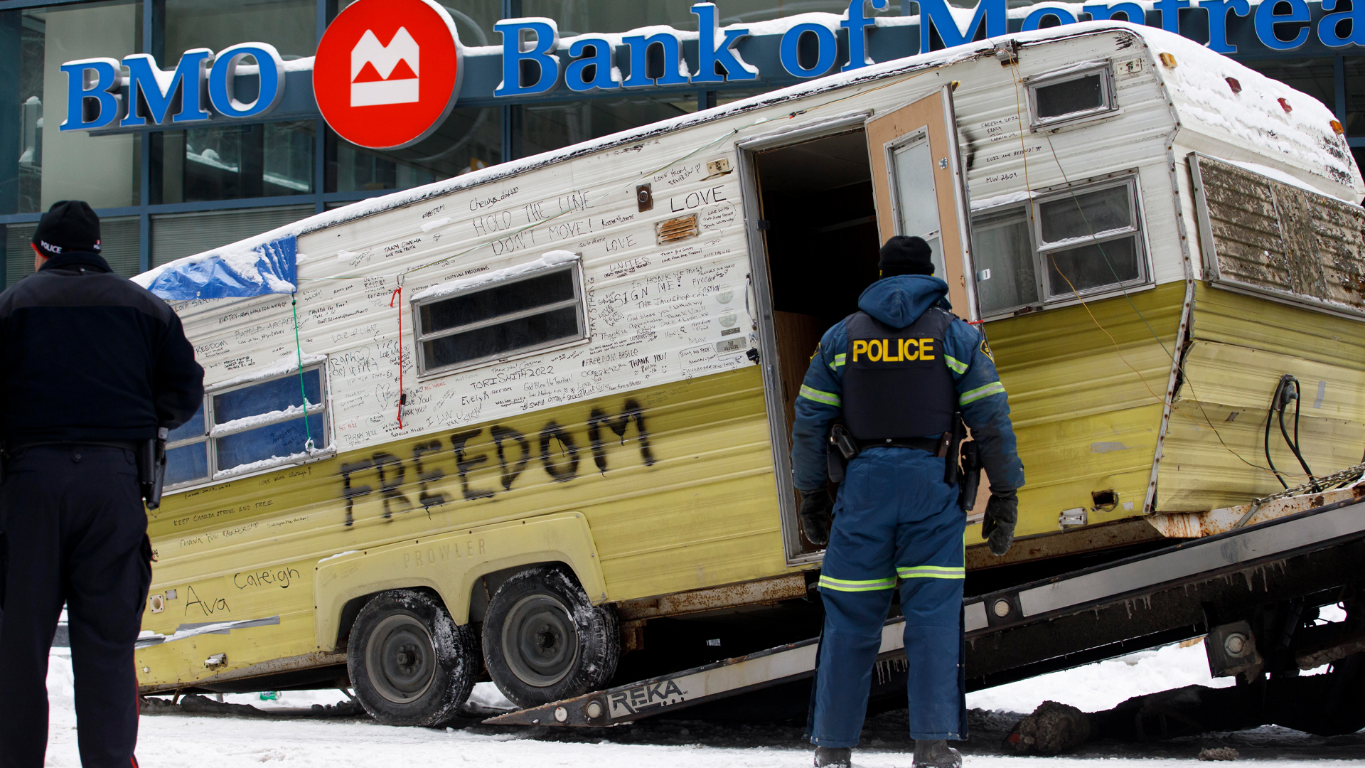 On a snow-covered street, a dingy-looking yellow and white camper covered with dozens of words like “freedom” is loaded onto a flatbed as two police officers supervise the operation.