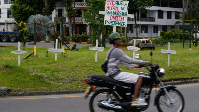 A young man drives a motorcycle past a small grassy area where the sign stands next to five white wooden crosses with one or two words on them. In the distance is a city block with apartment buildings in white stucco.