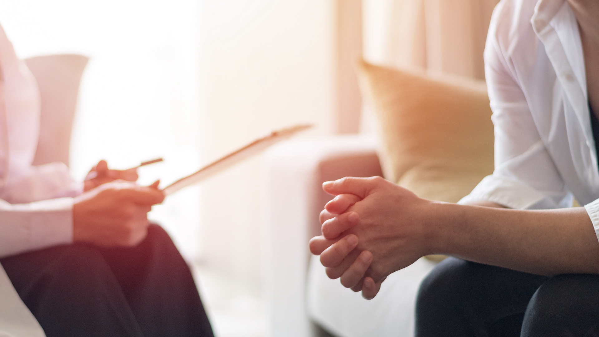 A woman sits on a sofa and leaning forward, hands clasped, speaking with a person sitting opposite her with a clipboard and pen in hand.