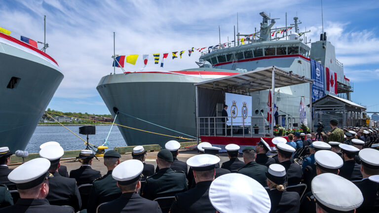 Two light-grey ships are tied up alongside a dock, each festooned with colourful nautical flags strewn from their bows to the top of their bridges. In front of them, a dais has been set up to face the crowd, which consists of four rows of seated navy personnel, all dressed in black uniforms with their flat-topped, white hats.