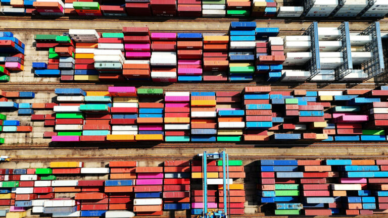 Hundreds of multi-coloured shipping containers arranged in dozens of straight lines are photographed from above.