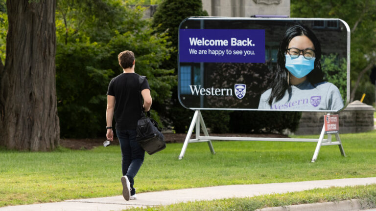 A student walks toward a Western University sign that says “Welcome back” featuring a woman wearing a blue surgical mask.