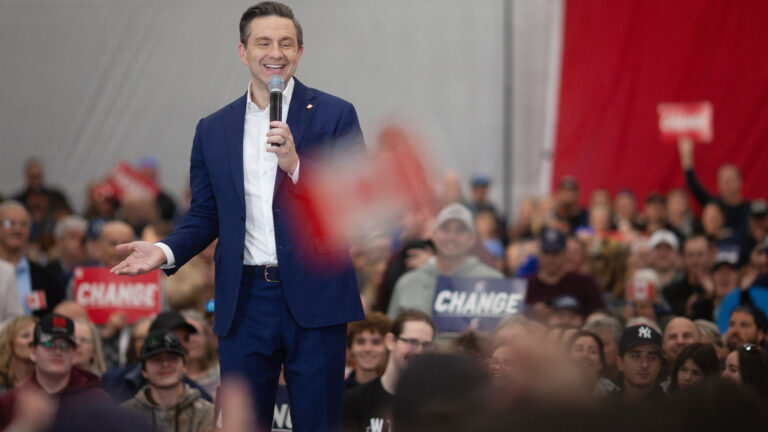 Poilievre is smiling as he stands in the middle of a room filled with seated people. A few of them hold signs saying “change.” Poilievre looks relaxed in a dark blue suit and white shirt. He has a mic to his mouth.
