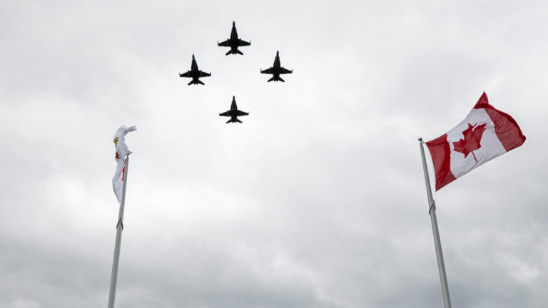 Four jets fly against a gray sky with a Canadian flag fluttering in the foreground.