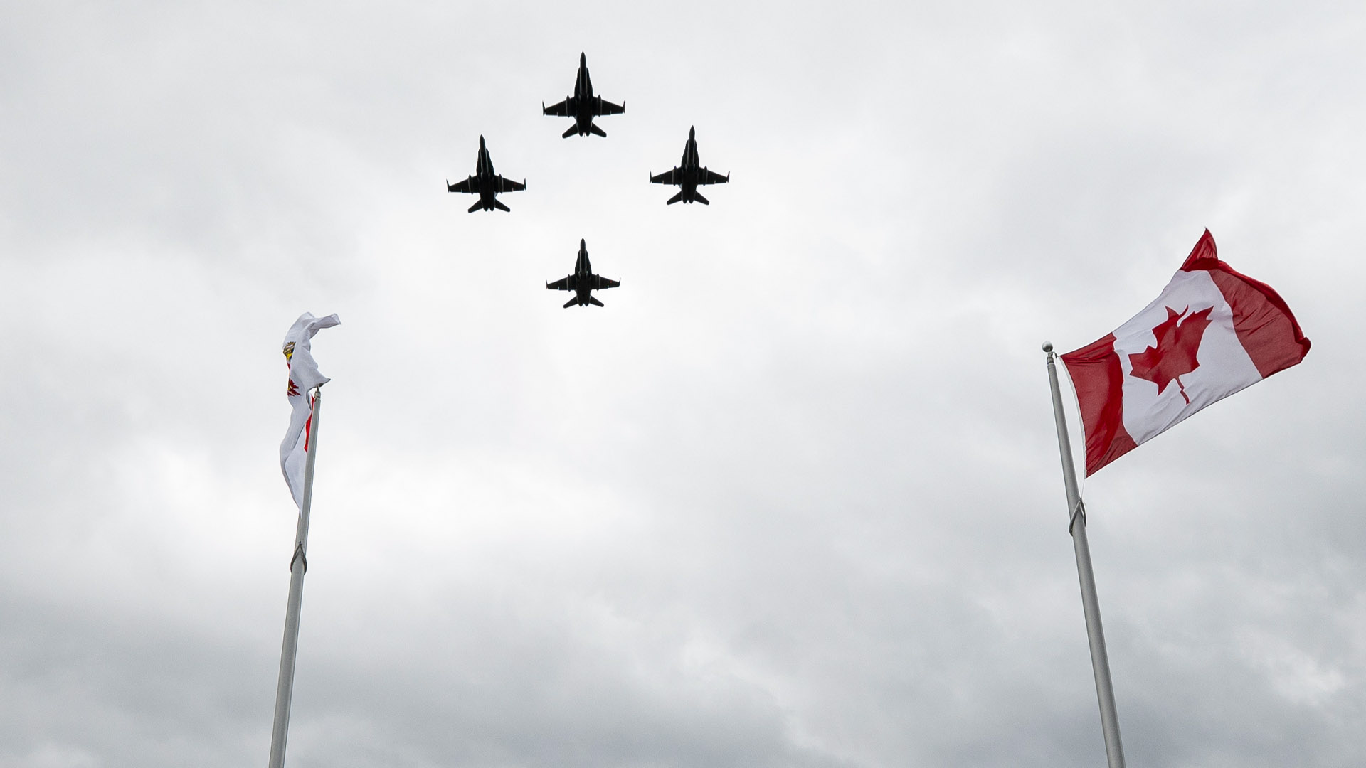 Four jets fly against a gray sky with a Canadian flag fluttering in the foreground.
