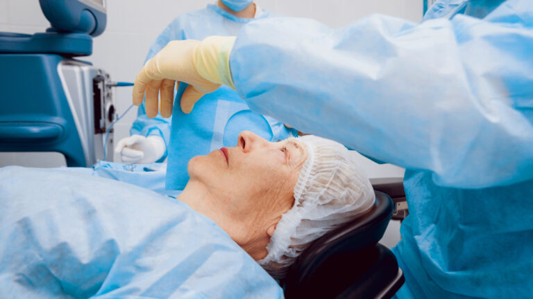 In a room with medical equipment, an older white patient is lying down, staring at the ceiling, with a sheer white net covering their hair. A person dressed in medical scrubs lifts a piece of blue fabric over one side of the patient’s face while another person in scrubs and mask stands nearby.