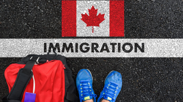 The word "immigration" is written on a white line that divides an asphalt surface in two. On one side, the Canadian flag, painted on the ground. On the other, a red travel bag and a pair of feet in blue sneakers.