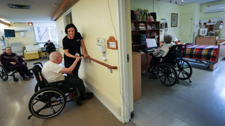 The worker is standing next to the wall in a hallway. She leans with one hand on a rounded wooden railing that runs the length of the wall at waist level. With her other hand, she lifts the hand of a man seated in a wheelchair facing the wall. He has a curve in his spine that bends him forward. Two other people watch from down the hallway. One is in a wheelchair, the other is seated under a window. The walls are pale yellow. The floor is brown linoleum.