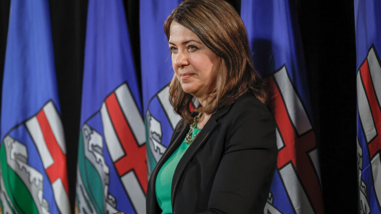 Smith stands in front of a row of large blue Alberta flags draped on their poles. She is holding a black plastic binder. She’s wearing a black jacket and a green shirt with a necklace.