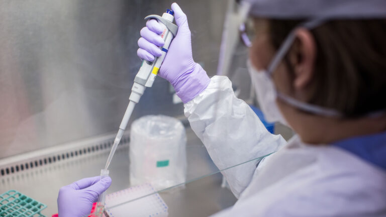 The technician is a woman who is directing a long, tapered tube into a small clear vial. She’s in a white protective covering with lilac-coloured rubber gloves.
