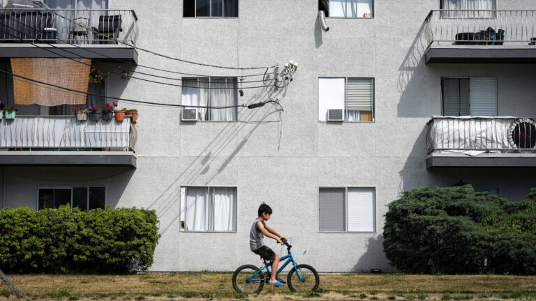 A young boy on a bike rides past a three-storey light-grey stucco apartment building with balconies. The windows are small and square. Hedges line the first floor.