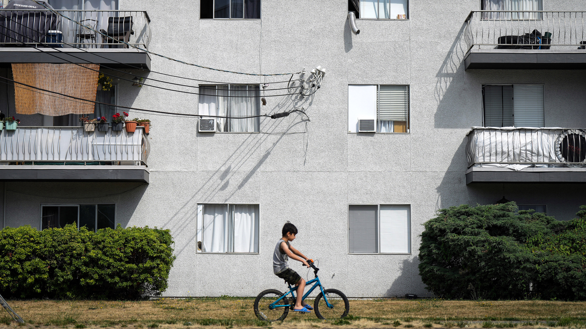 A young boy on a bike rides past a three-storey light-grey stucco apartment building with balconies. The windows are small and square. Hedges line the first floor.