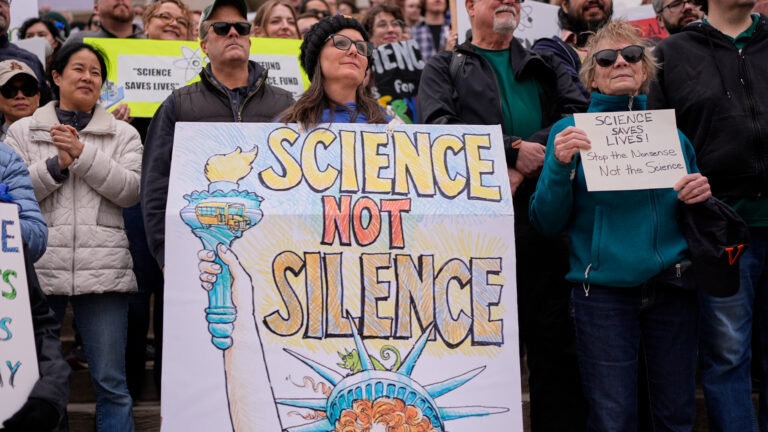 Protesters stand behind a banner with a hand-drawn Statue of Liberty and with the slogan “Science not silence” written on it.