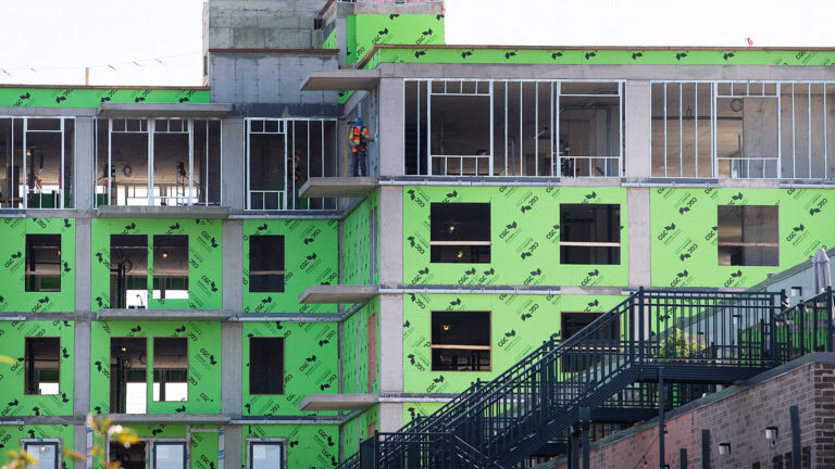A low-rise condo project of about four storeys is partially built, with exposed concrete and lime-green membranes covering most of the surfaces. The spaces for window have no glass yet. Two men work on the roof and top floor.