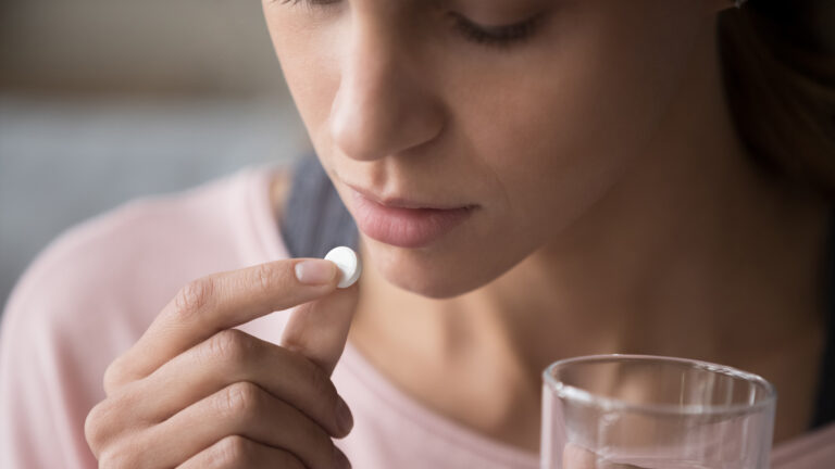Young white woman holds a round, white pill to her mouth, with a glass of water in her other hand.