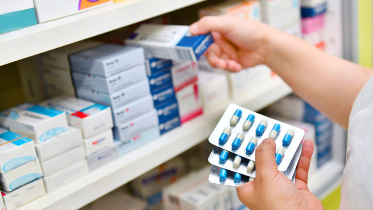 The hands of a pharmacist taking a medicine box and capsule pack from the shelves of a pharmacy.