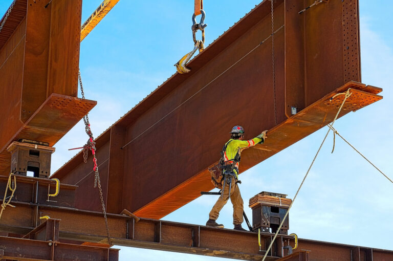 A worker guides the placement of a huge steel girder suspended by a crane. He is standing on a smaller length of steel high in the sky.