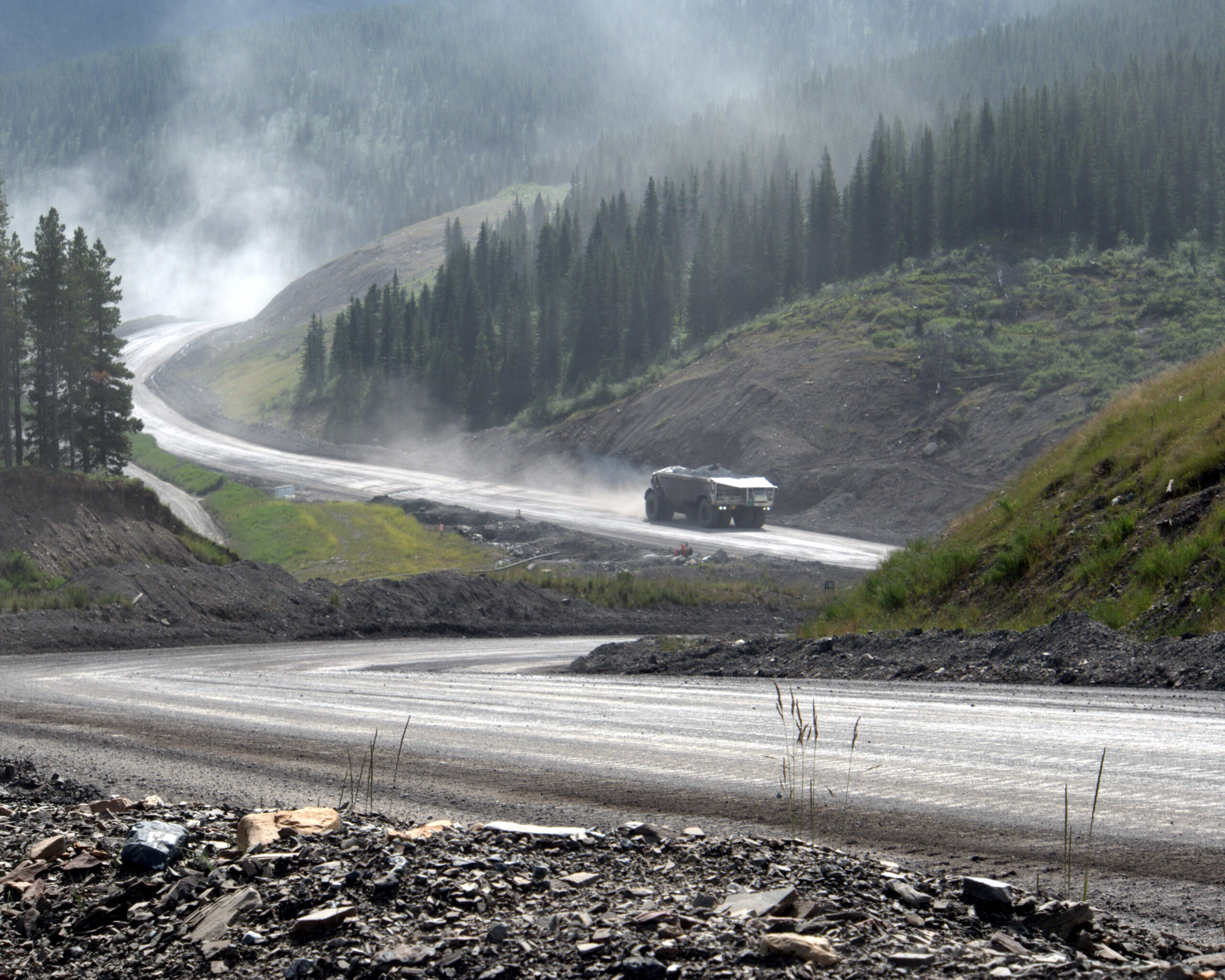 The truck kicks up dust as it travels a working road with no markings. The road cuts through large swaths of untouched forest as well as areas that have been cleared.