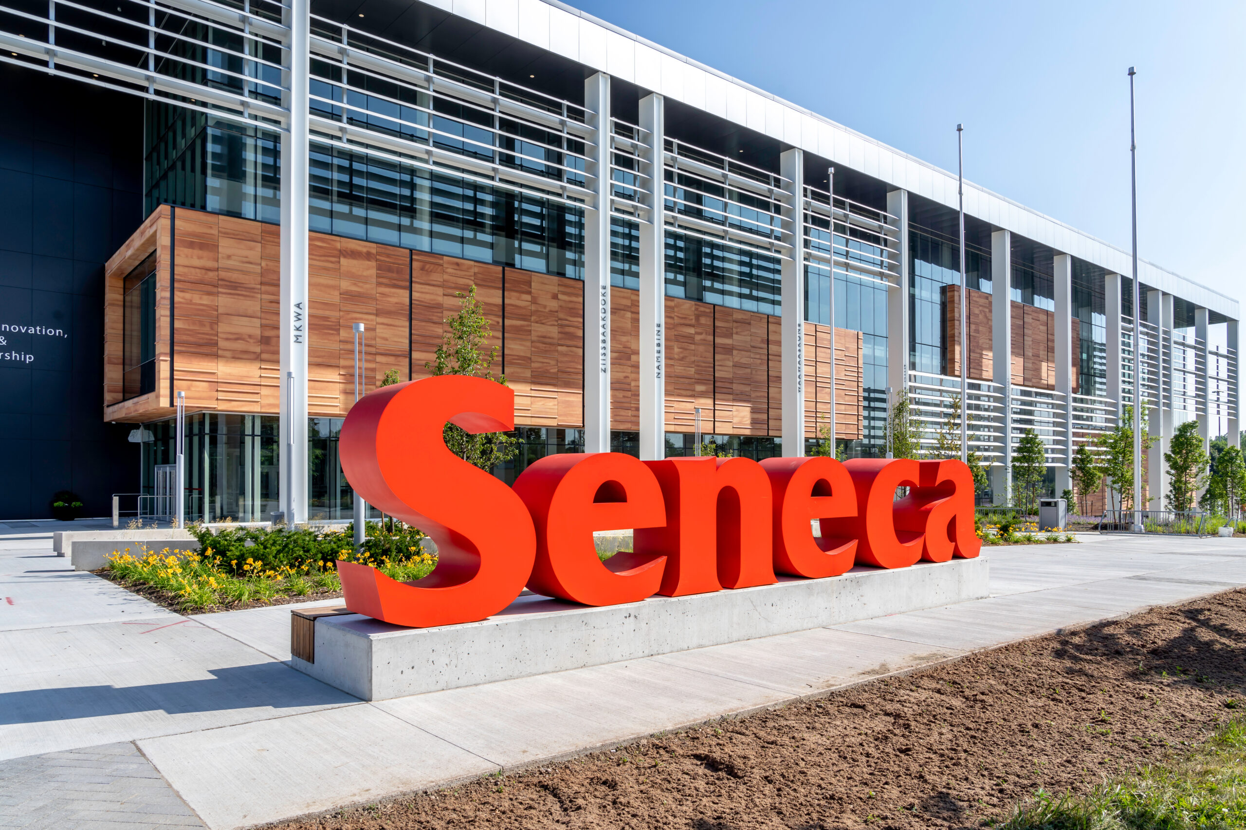 A prominent and bright red sign made up of the word “Seneca” sits in front of a long contemporary building with glass and accents of wood panel and thin white metal bars placed here and there.