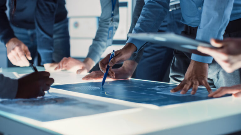 Five men lean on a light table, making calculations with pens and diagrams.